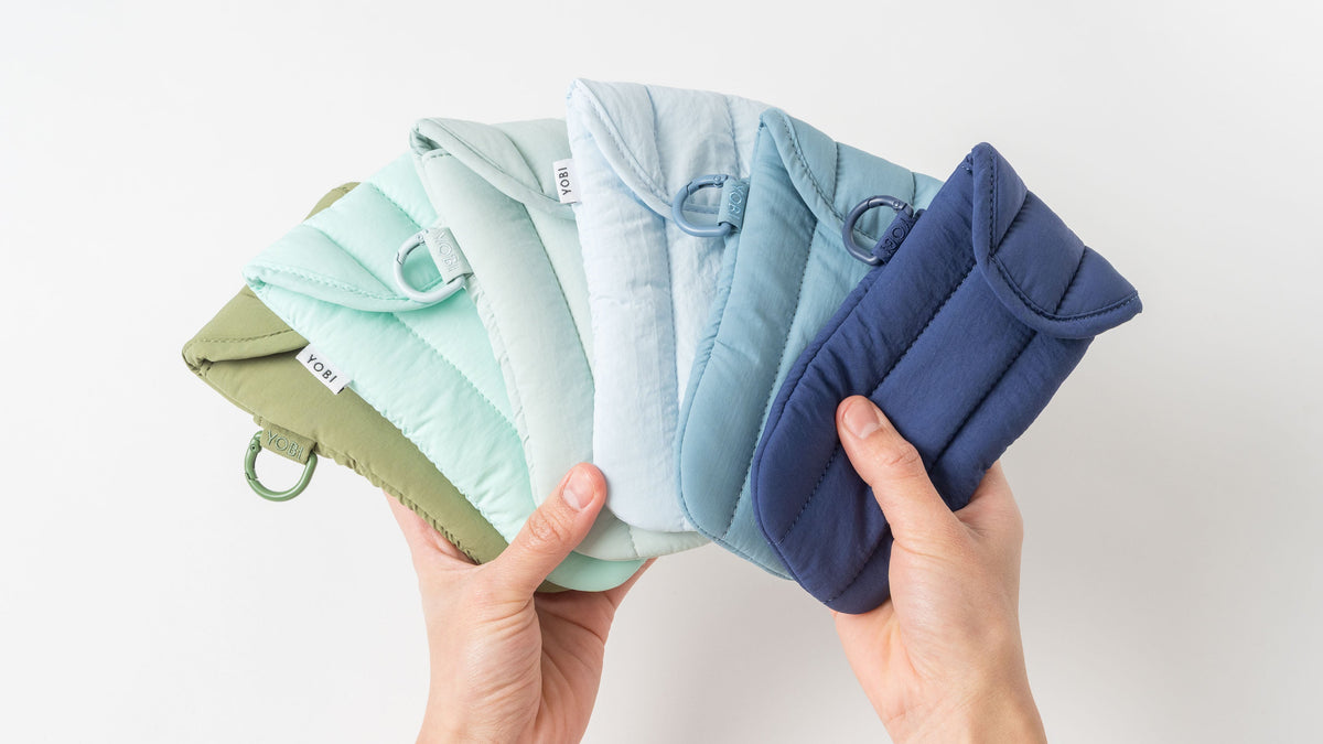 A pair of hands holding a fan of quilted fabric glasses cases in multiple soft colors, including sage green, mint, light blue, medium blue, and navy, against a clean white background.