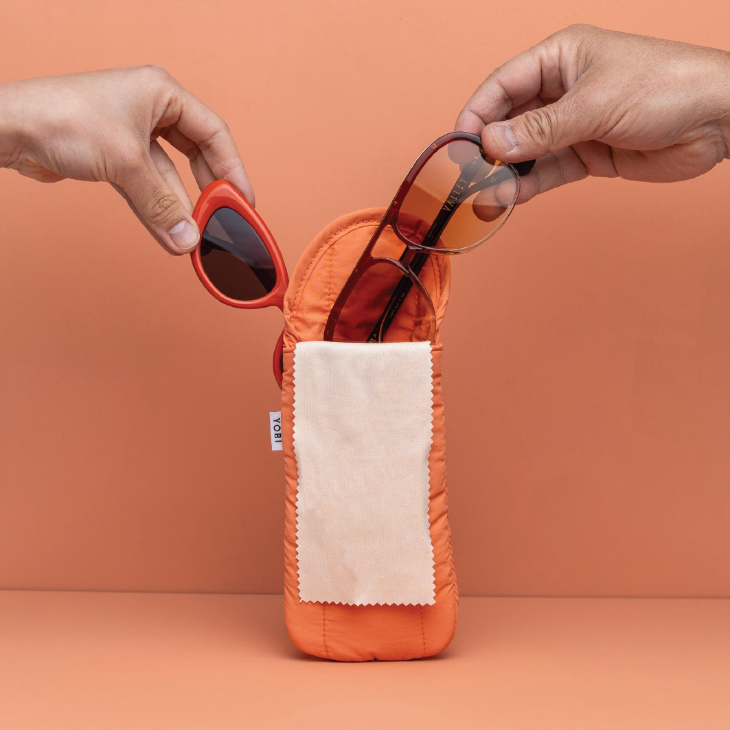 Orange sunglasses case with sunglasses being placed into it against a peach background